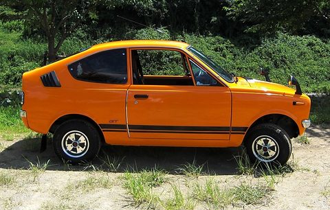 Side view of an orange small coupé with black stripes, parked on dusty ground with weeds. Opens a larger view
