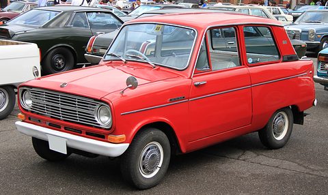 Bright red small saloon with a chrome grille and round headlamps, other cars behind. Opens a larger view