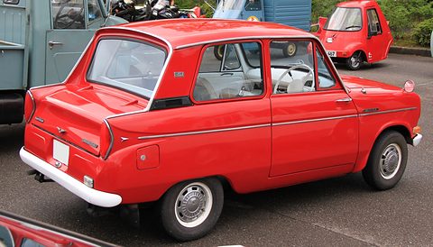 Rear three-quarter of a red small saloon with chrome trim, a motorcycle and a three-wheeler behind. Opens a larger view