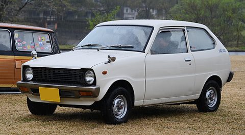 White small hatchback with a black grille on dry grass, a wood-panelled car behind. Opens a larger view