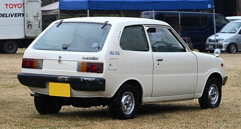 Rear three-quarter of a white small hatchback on dry grass, a delivery truck behind. Opens a larger view