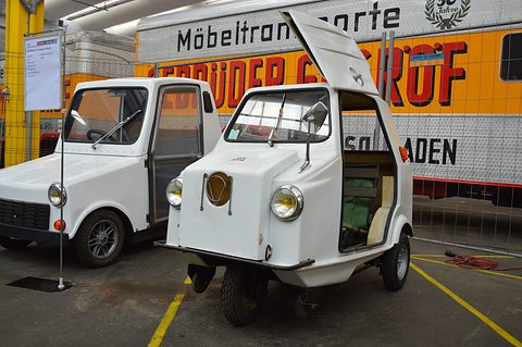 White three-wheeler with its upper door hinged up showing green seats, next to a white four-wheeled microcar and a removals lorry.