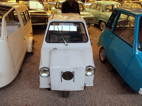 Front view of a narrow white three-wheeler with a square central cowl carrying a round grille, headlamps at either side. Opens a larger view