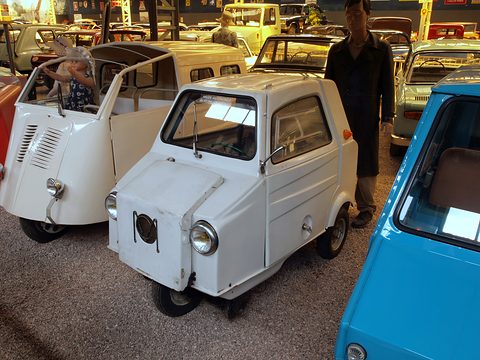 Two small white three-wheelers in a museum hall, the left one with its side door folded up, a figure in a dark coat behind.
