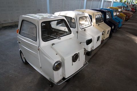 Line of small three-wheelers, white ones in front and blue, black and yellow ones behind, under a long shed roof.
