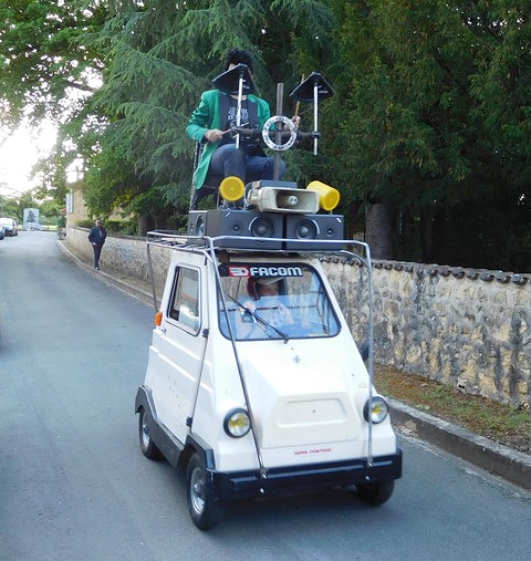 A small white boxy microcar with a frame on its roof holding speakers and a costumed performer, on a village road. Opens a larger view