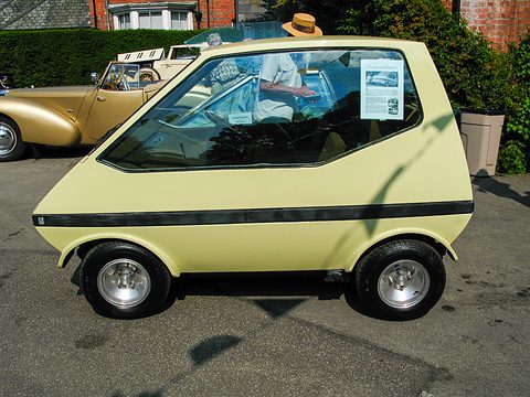 Cream city car side-on with a long sloping windscreen and no side doors, vintage cars behind.