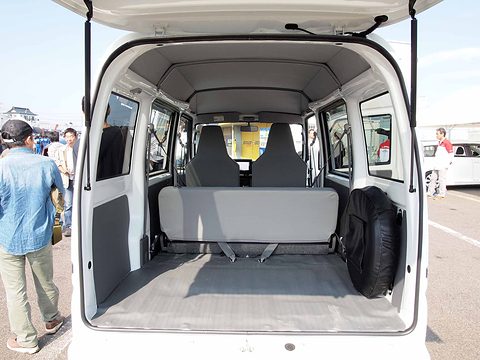 Rear view into a white van’s empty load area with folded rear seats, tailgate raised, people standing beside. Opens a larger view