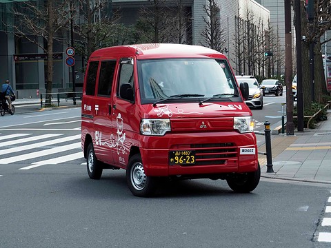 Red tall kei van with white cartoon bear graphics driving on a city street by a pedestrian crossing.