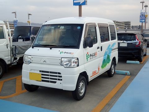 White tall kei van with green and blue livery and a yellow plate in a car park marked in blue and orange. Opens a larger view