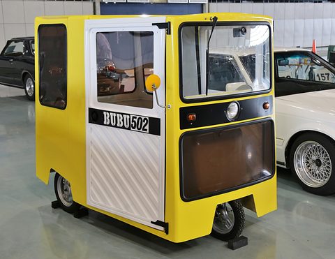 Boxy yellow and white single-seat microcar with large windows and a BUBU 502 panel on its door, on a hall floor.