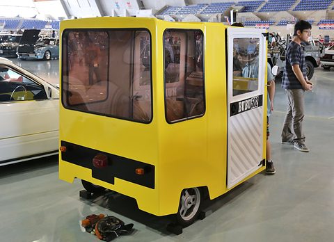 Rear quarter of a boxy yellow single-seat microcar with large windows, a visitor standing beside it in an exhibition hall. Opens a larger view