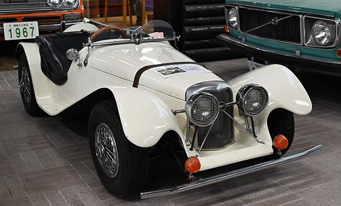 Small white open roadster with sweeping wings, twin round headlamps and a mesh grille, among classic cars indoors. Opens a larger view
