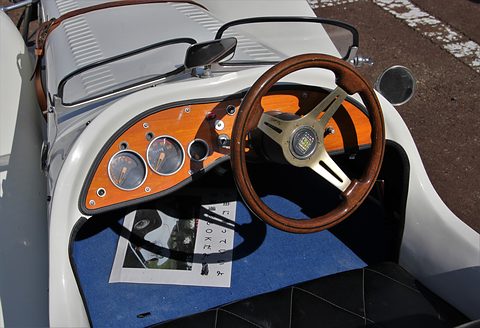 Open cockpit with a wood-rimmed steering wheel, orange dashboard with round dials and a blue floor mat. Opens a larger view