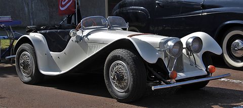 Side view of a small white open roadster with long flowing wings, wire wheels and twin headlamps, parked indoors. Opens a larger view