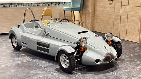 Silver open single-seat microcar with a vertical-bar grille, cycle wings and a roll hoop on a stone floor. Opens a larger view