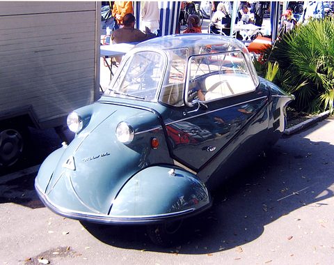 Blue-grey bubble car seen from the front quarter, palms and visitors behind. Opens a larger view