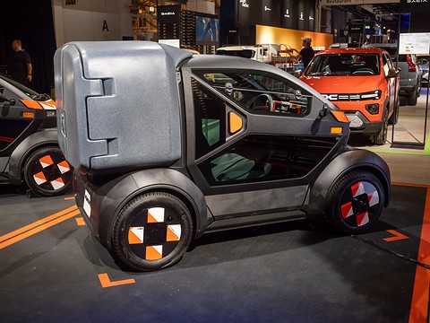 Grey narrow two-seat electric car with a large cargo box behind the cabin and orange-patterned wheels on a stand.