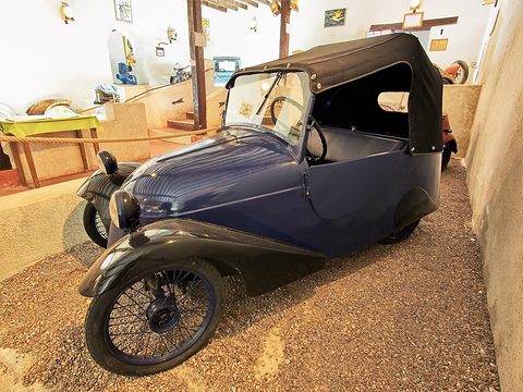 Dark blue two-seater with a black hood and spoked wheels in a museum bay with tools on the wall. Opens a larger view