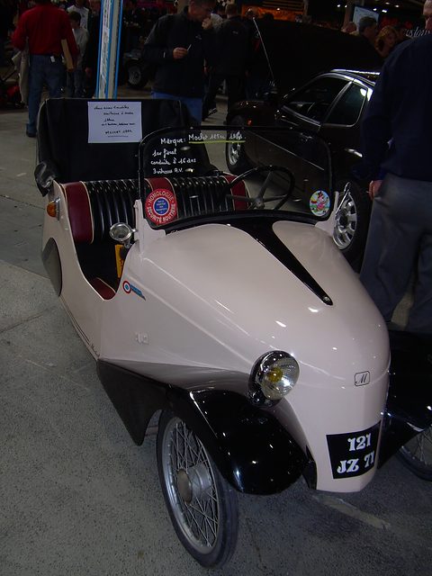Pale pink open two-seater with black wings and red seats, a sign board on its windscreen. Opens a larger view