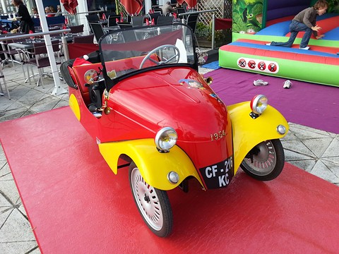 Red open two-seater with yellow wings on a red mat, a bouncy castle behind. Opens a larger view