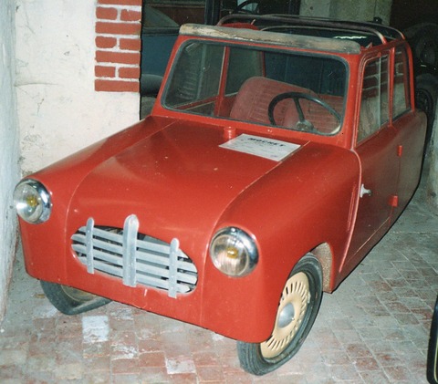 Faded red small coupé with grey grille bars and cream wheels in a whitewashed building. Opens a larger view