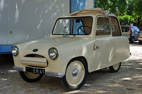 Cream small two-seat coupé with a folded fabric roof, parked on gravel in dappled shade.