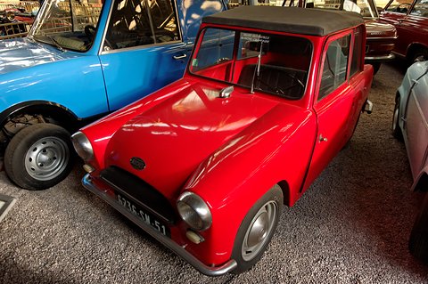 Red small two-seat car with a grey fabric roof on a gravel museum floor beside a blue car. Opens a larger view