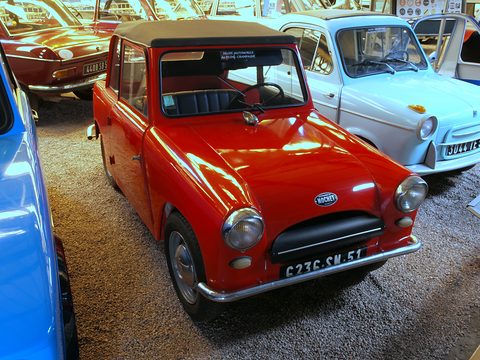Red small two-seat car with a fabric roof and chrome bumper, packed among other small cars. Opens a larger view