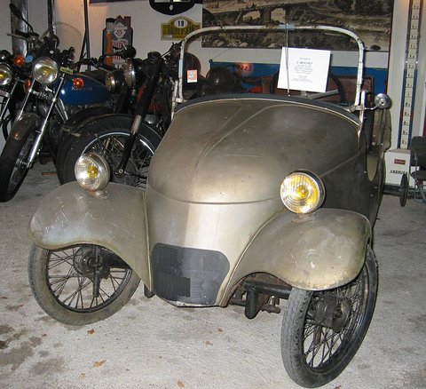 Weathered grey small open car with cycle wings and spoked wheels, motorcycles all around it. Opens a larger view