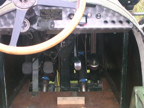A wooden steering wheel before a metal dash carrying a maker’s plate, two pedal cranks in the footwell below. Opens a larger view
