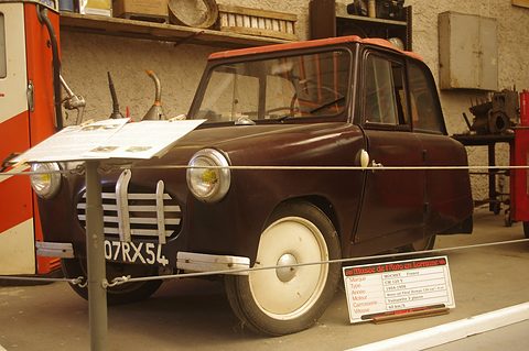 Black small coupé with a slatted grille and cream wheels behind a rope with a museum sign. Opens a larger view