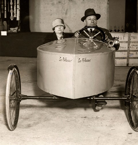 Black-and-white: a man in a bowler hat and a woman in a cloche hat seated in a boxy pedal car.
