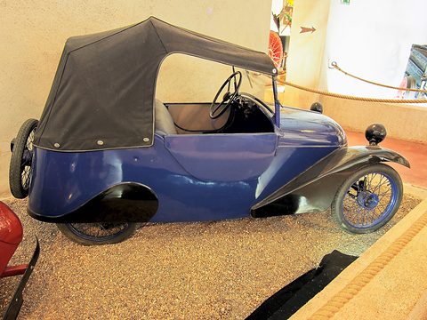 Dark blue open two-seater with black cycle wings and a black hood on a gravel display bed.