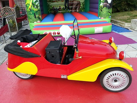Side view of a red and yellow open two-seater on a red mat beside an inflatable play castle. Opens a larger view