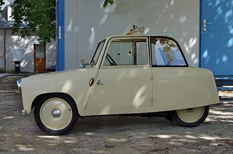 Side view of a cream two-seat coupé with cream wheels in front of a blue shed. Opens a larger view