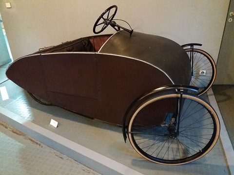 Side view of a long dark plywood shell on four bicycle wheels with a small steering wheel on a column. Opens a larger view
