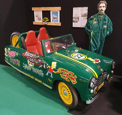 Green open two-seater with stickers and yellow wheels beside a life-size cut-out of a driver. Opens a larger view