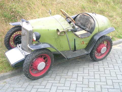 Green pedal car shaped like a vintage sports car with red wire wheels, on brick paving. Opens a larger view