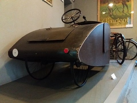 A dark varnished plywood pedal car with a rounded tail and a steering wheel, a Peugeot poster on the wall behind. Opens a larger view