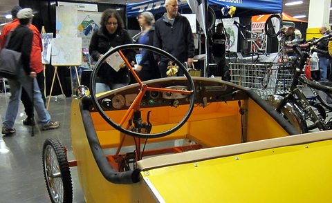 Looking into a yellow pedal car: an orange-spoked steering wheel, a bench seat and two sets of pedals.