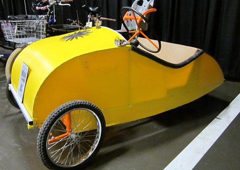 A bright yellow pointed pedal car on spoked bicycle wheels with an orange steering wheel, bicycles behind. Opens a larger view