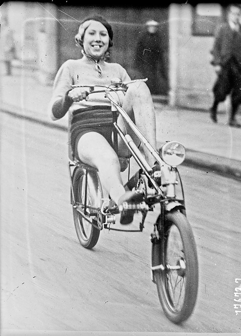 A smiling woman in a jumper and shorts pedalling a low recumbent bicycle with a front lamp along a street.