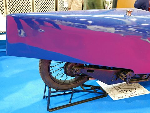 A spoked rear wheel and drive chain beneath a blue pointed tail, the car resting on a metal stand. Opens a larger view
