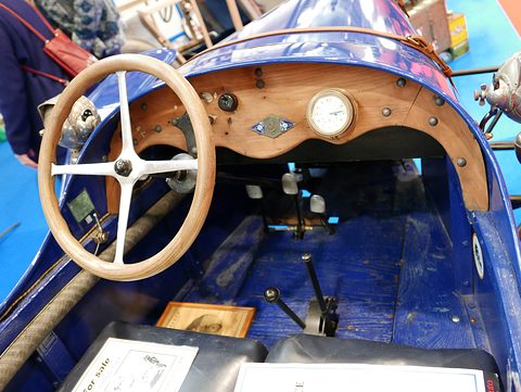 A wooden dashboard with a single round gauge, a wood-rimmed steering wheel and a gear lever on a blue floor. Opens a larger view