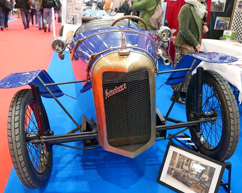 Front view of a narrow blue car with a script radiator, one front wheel and small wheels on outriggers to each side. Opens a larger view