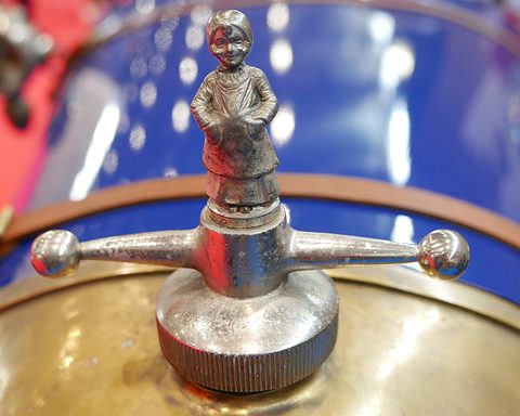 A small silver figure of a child standing on a winged radiator cap above blue paintwork. Opens a larger view