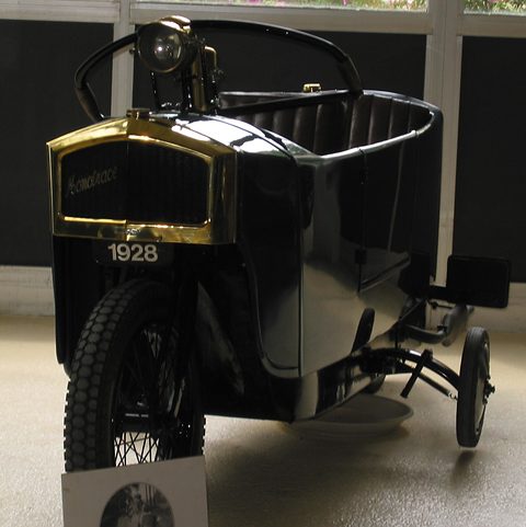 A narrow black car with a brass radiator, a single headlamp on a stalk and an outrigger wheel at its side. Opens a larger view