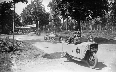 A narrow white single-track car with two helmeted men aboard rounds a dusty woodland bend, another small car behind. Opens a larger view