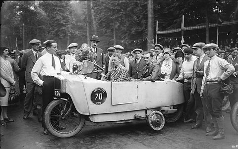 A long white narrow car with a race number disc, two men seated in tandem, surrounded by a crowd in caps and hats. Opens a larger view